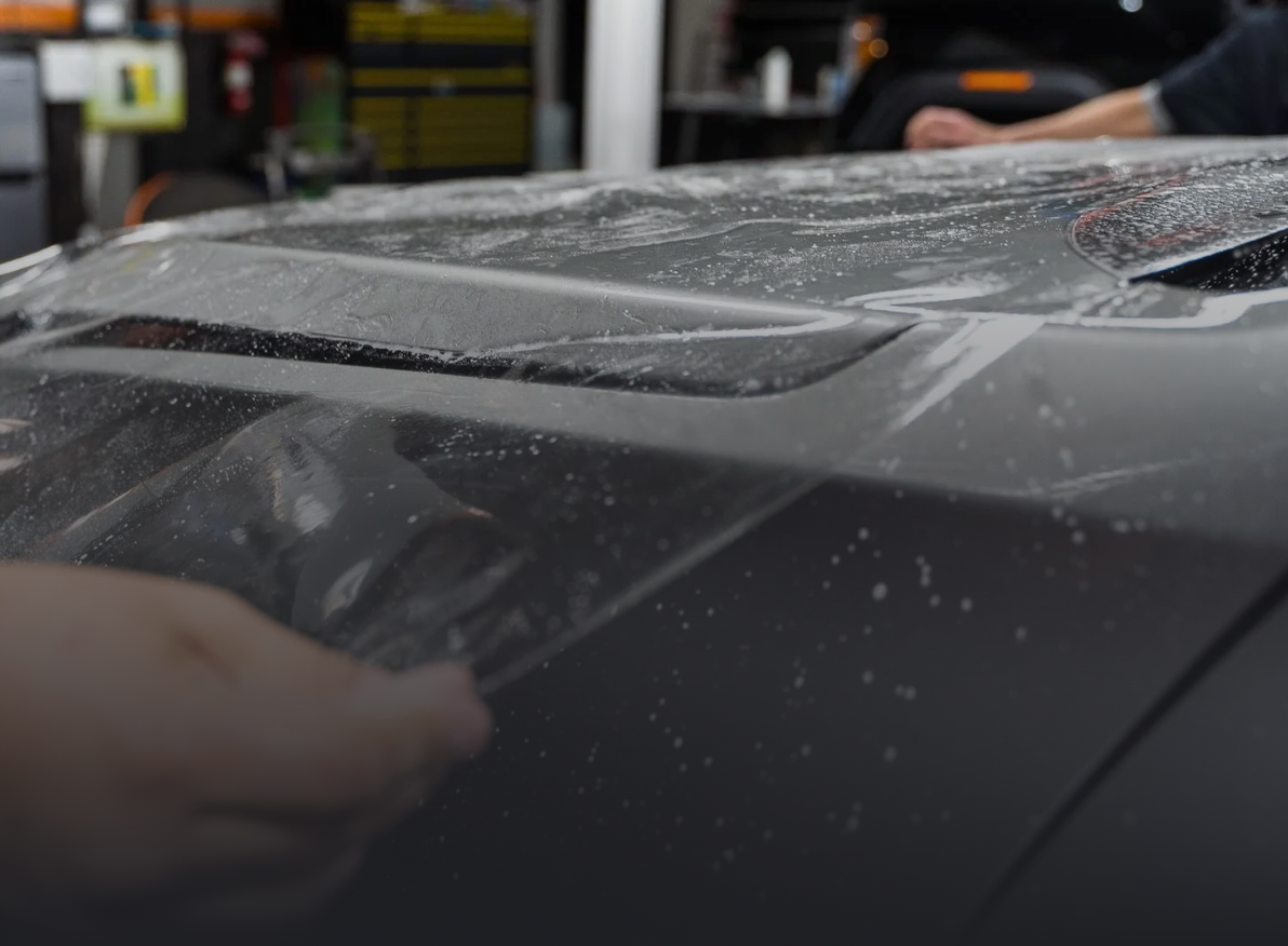 Person applying clear protective film to a car's hood in a shop.
