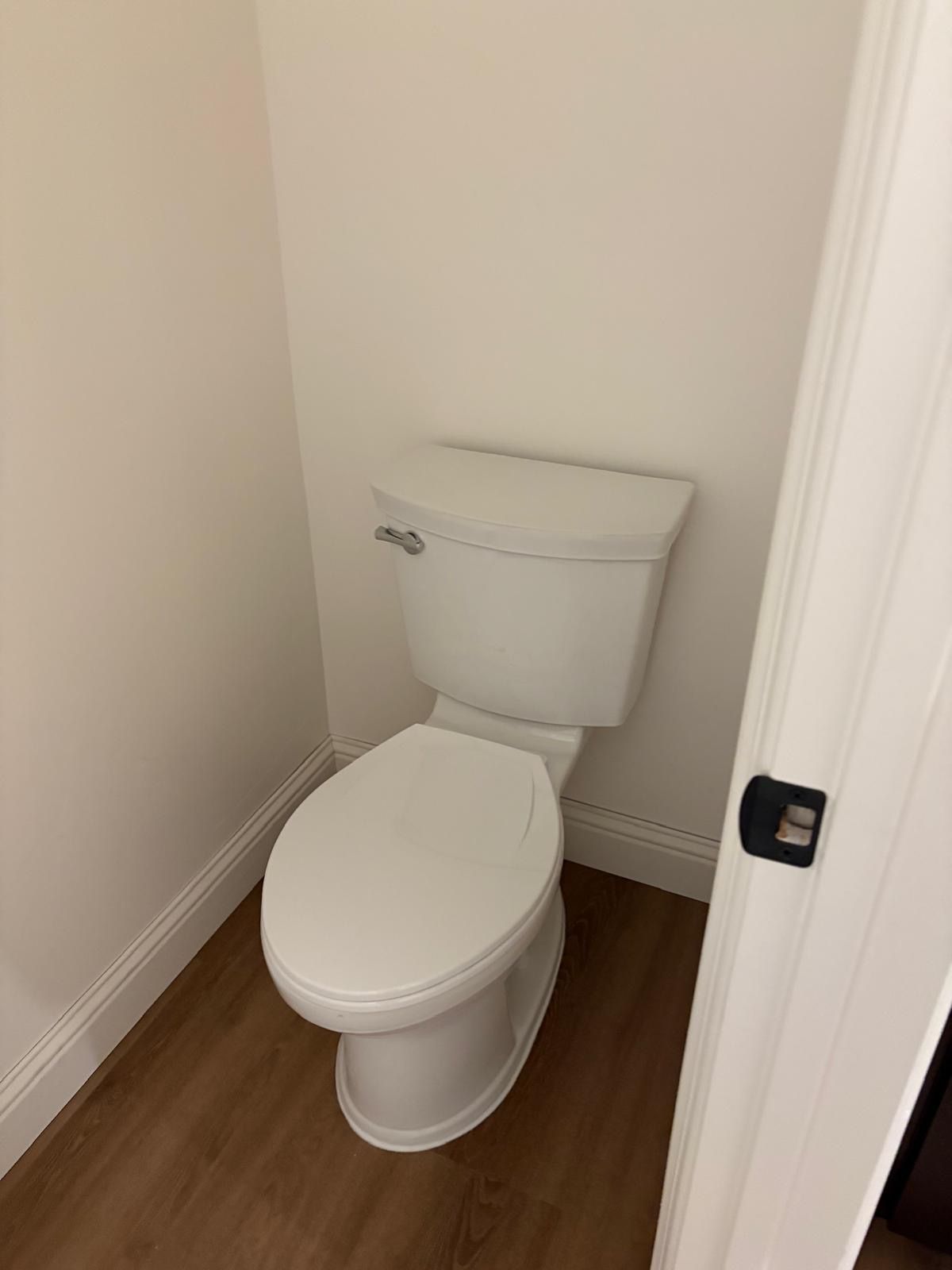 White toilet in a small bathroom with wood-look flooring and white walls.