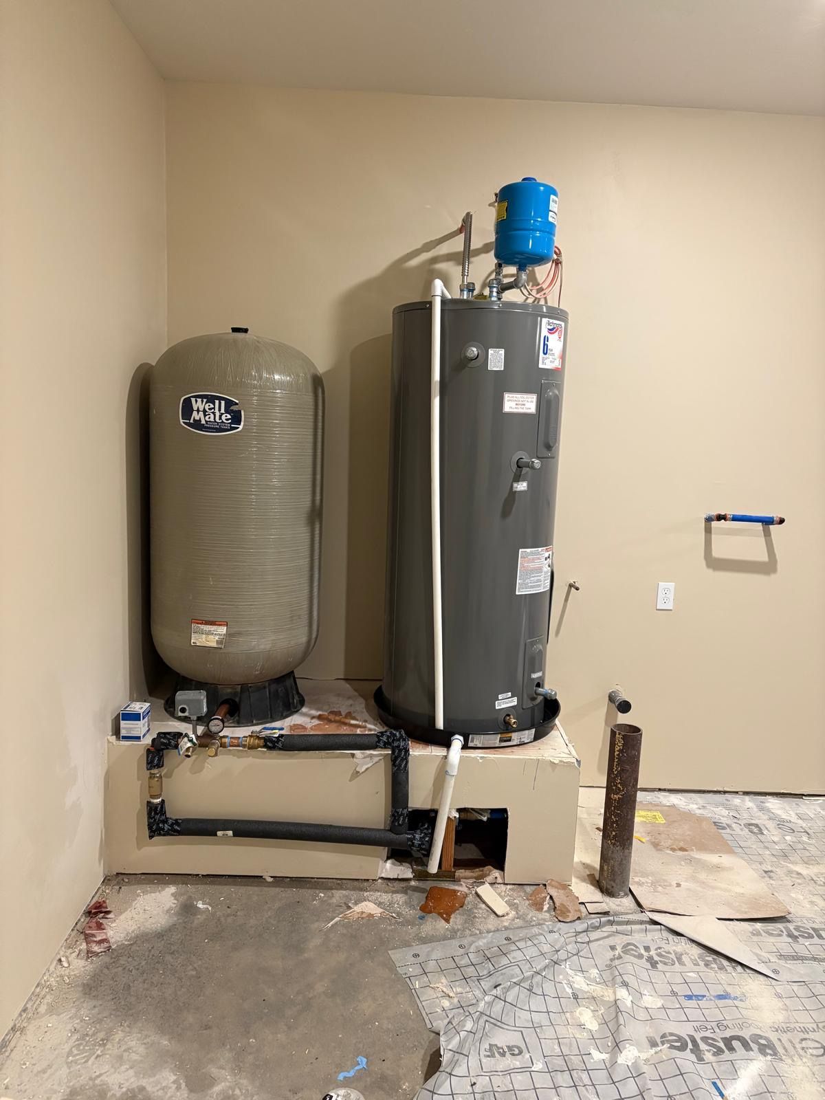 Water heater and pressure tank in a room under construction with exposed flooring and plumbing.