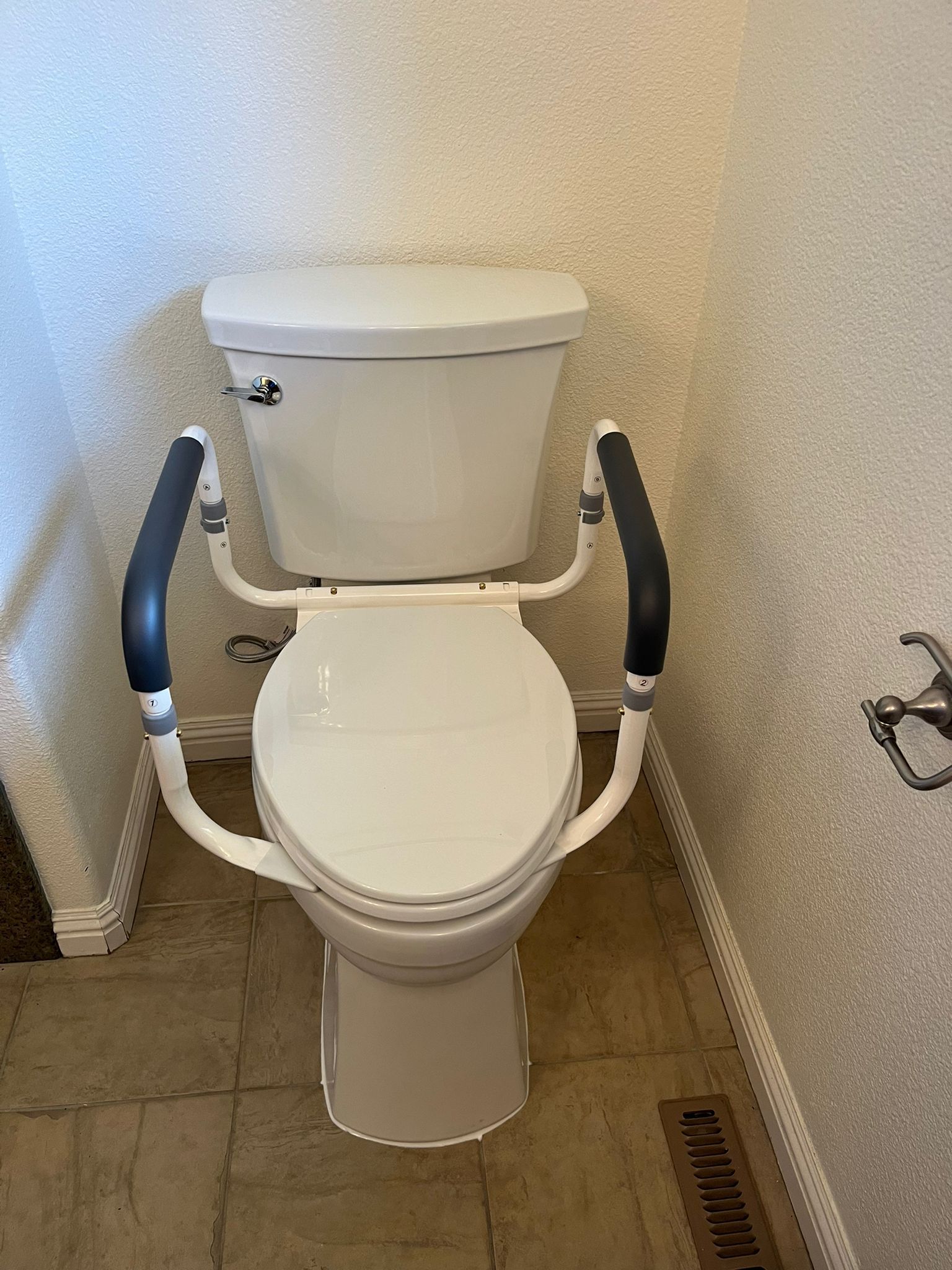 White toilet with safety grab bars, in a small bathroom with light-colored tile and textured walls.
