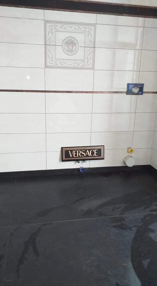 In una stanza, la parete è rivestita di piastrelle bianche con una piastrella recante l'emblema di Versace, la targa con il logo di Versace e il pavimento è scuro.