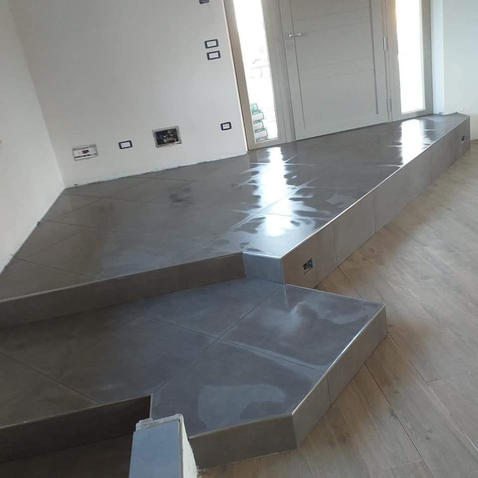 Una piattaforma rialzata rivestita di piastrelle grigie, con un gradino, addossata a una parete bianca con prese elettriche, adiacente a un pavimento in legno.