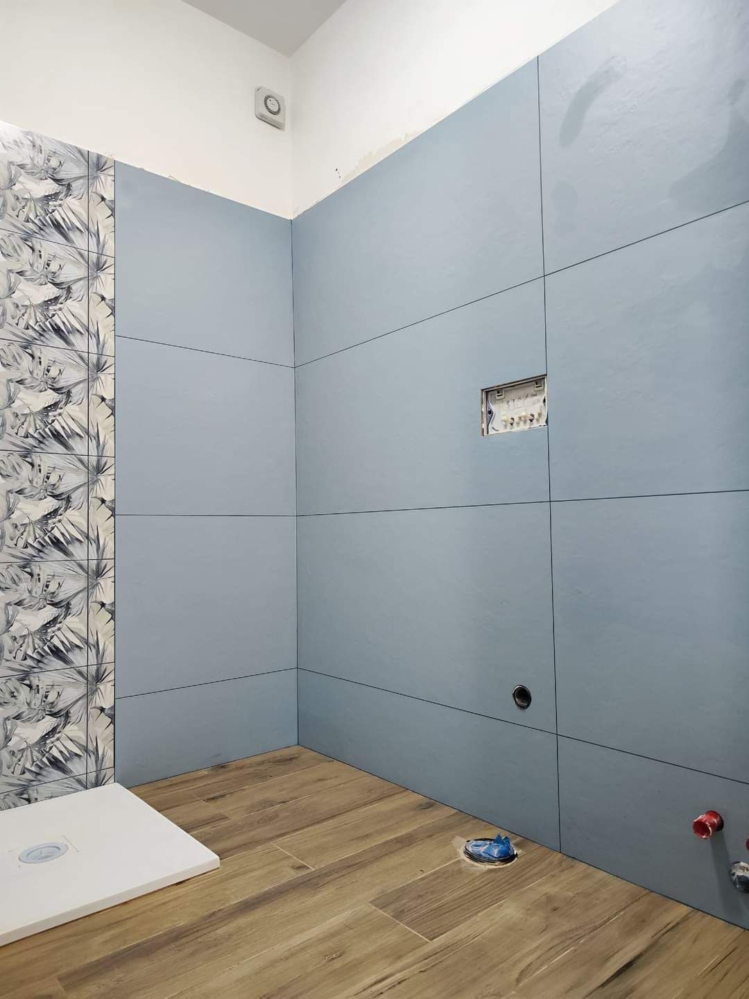 Un bagno in fase di ristrutturazione con pareti rivestite di piastrelle grigio-blu, pavimento effetto legno e una parete decorativa con motivi decorativi.