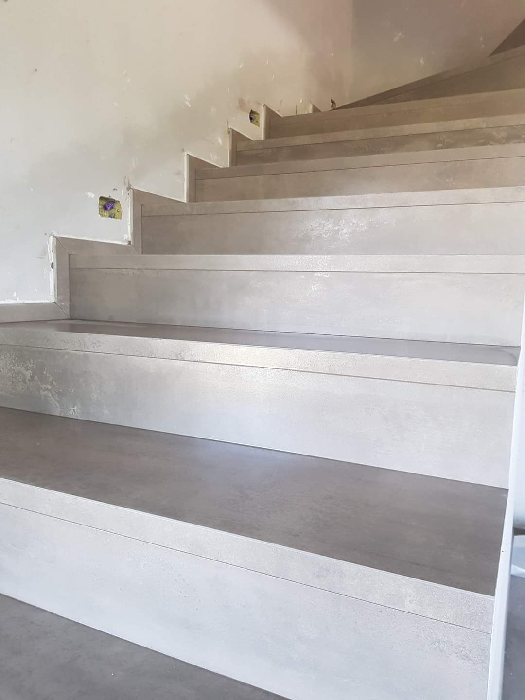 Una scala interna non rifinita con gradini in cemento e alzate verticali di colore chiaro che si stagliano contro una parete bianca.