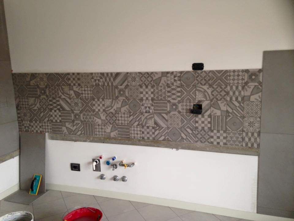 Un paraschizzi da cucina con piastrelle grigie decorate su pareti bianche, tubature a vista e piastrelle grigie sul pavimento.