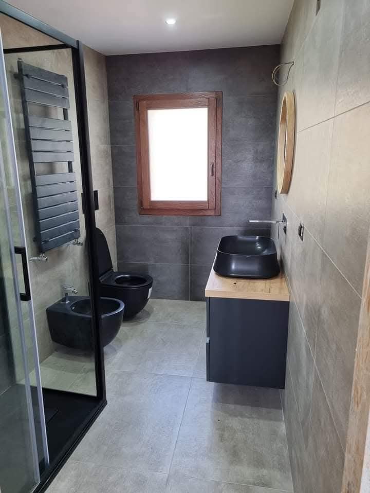 Bagno moderno con pareti rivestite di piastrelle grigie, doccia nera, lavabo a parete, bidet, WC e finestra.