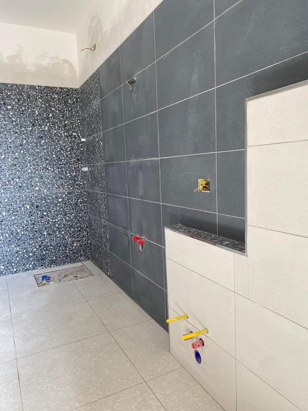 Un bagno in fase di ristrutturazione caratterizzato da un pavimento in stile terrazzo, una parete rivestita con piastrelle grigio scuro e una parete non rifinita con piastrelle bianche.