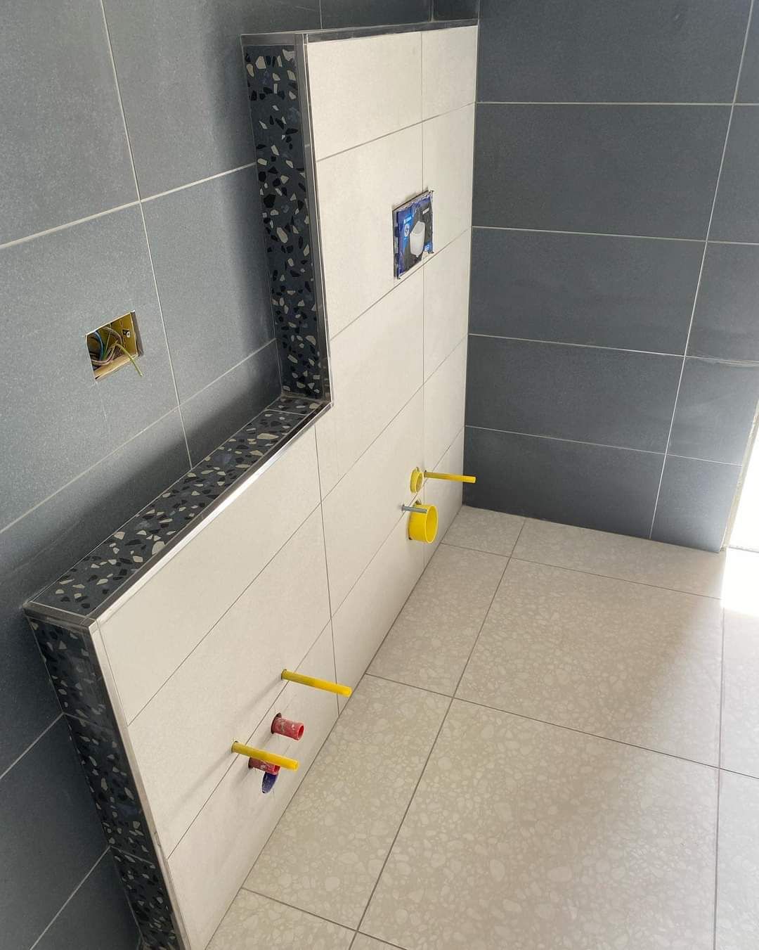 Una parete in fase di piastrellatura in un bagno, con una sezione di mensole rialzate e rubinetteria che sporge dalle piastrelle bianche.