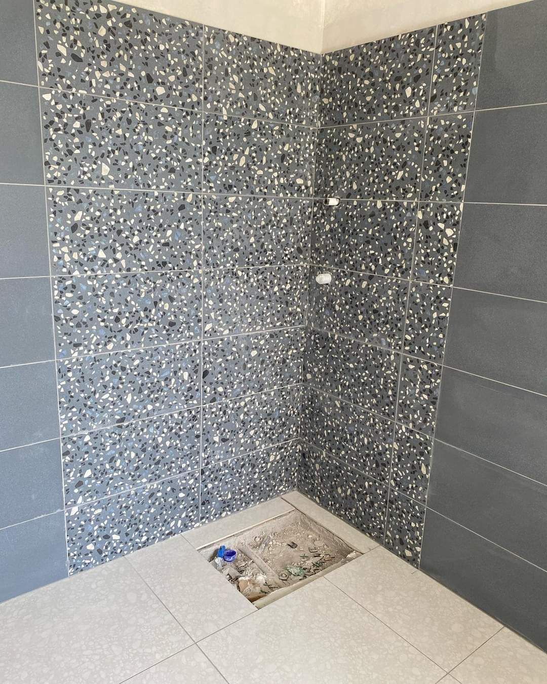 Un box doccia angolare con rivestimento a parete in terrazzo grigio antracite, piastrelle laterali grigie a tinta unita e piastrelle per il pavimento di colore chiaro.