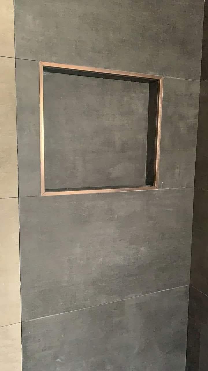 Una nicchia quadrata a muro con finiture in bronzo, incastonata nella parete del bagno rivestita di piastrelle grigio scuro.