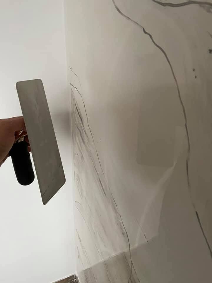 Una mano impugna una cazzuola di metallo contro una parete rivestita di piastrelle con motivo marmoreo bianco e grigio.