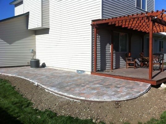 A backyard patio made of multi-colored pavers, partially covered by a wooden pergola attached to a tan-sided house.