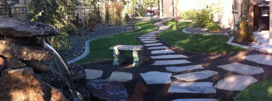 A stone pathway winds through a grassy yard featuring a stone fountain and a decorative stone bench.