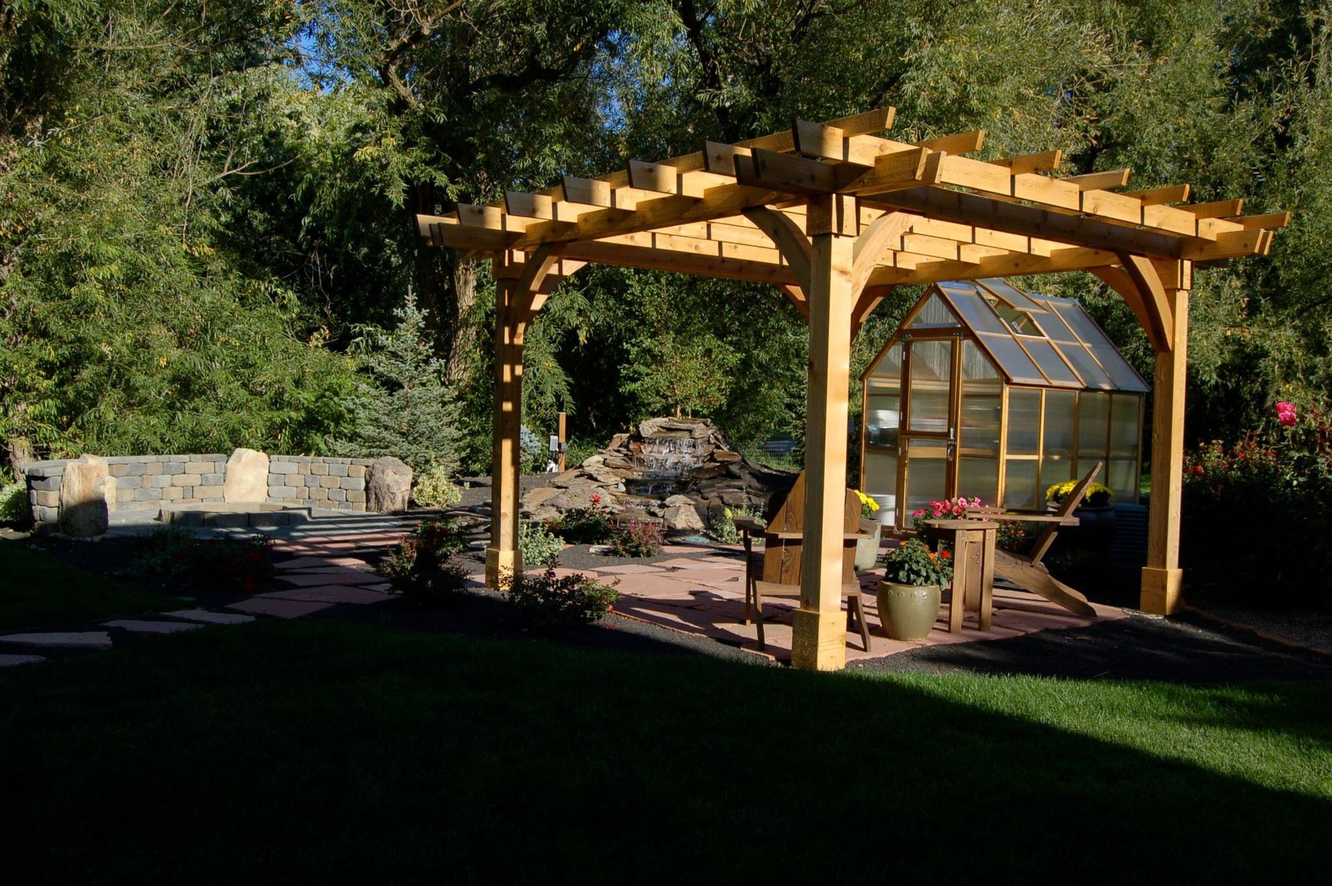 A wooden pergola sits over a patio with a small greenhouse and stone rock feature in a lush garden setting.