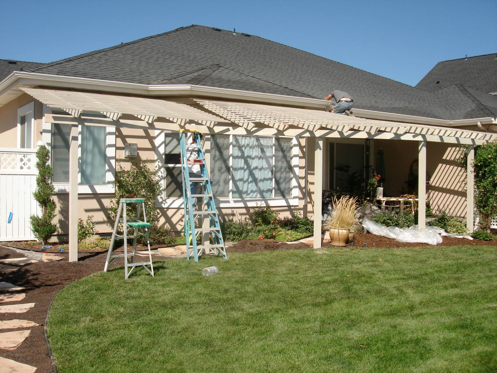 A worker on a ladder installs a light-colored wooden pergola attached to the rear of a residential house.