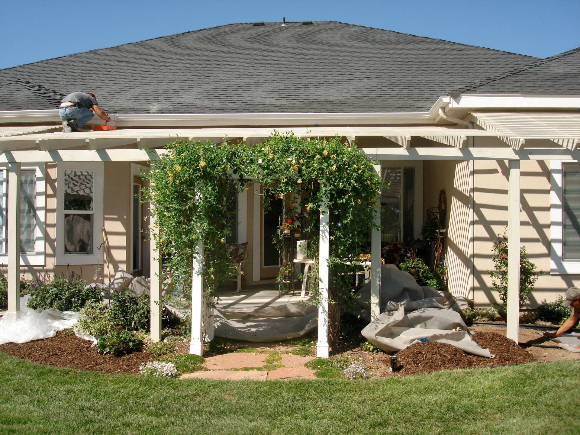A person works on the roof above a tan pergola covered in climbing vines, located at the back of a house with a lawn.