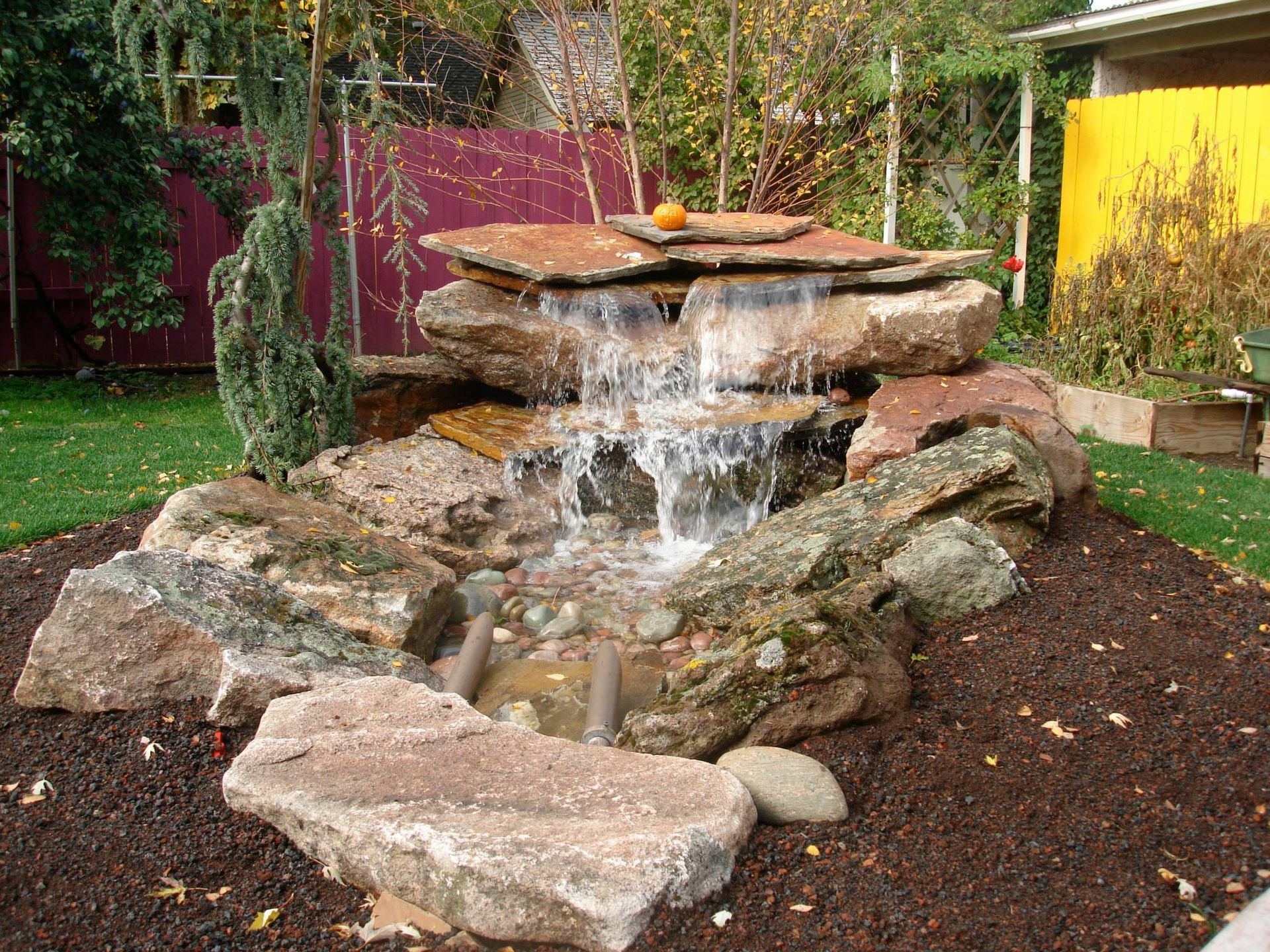 A tiered stone waterfall feature sits in a landscaped garden bed with dark mulch, surrounded by grass and foliage.