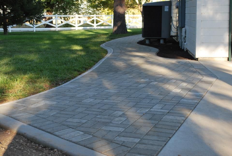 A gray paver walkway curves along a lawn and a white building exterior on a sunny day.