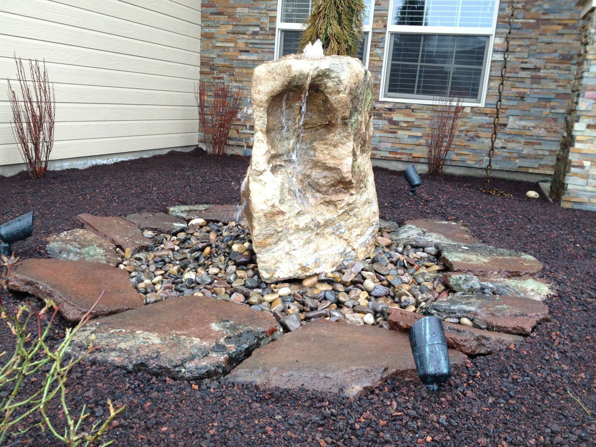 A stone fountain stands in a garden bed with dark mulch, surrounded by flat rocks and small landscape lights.
