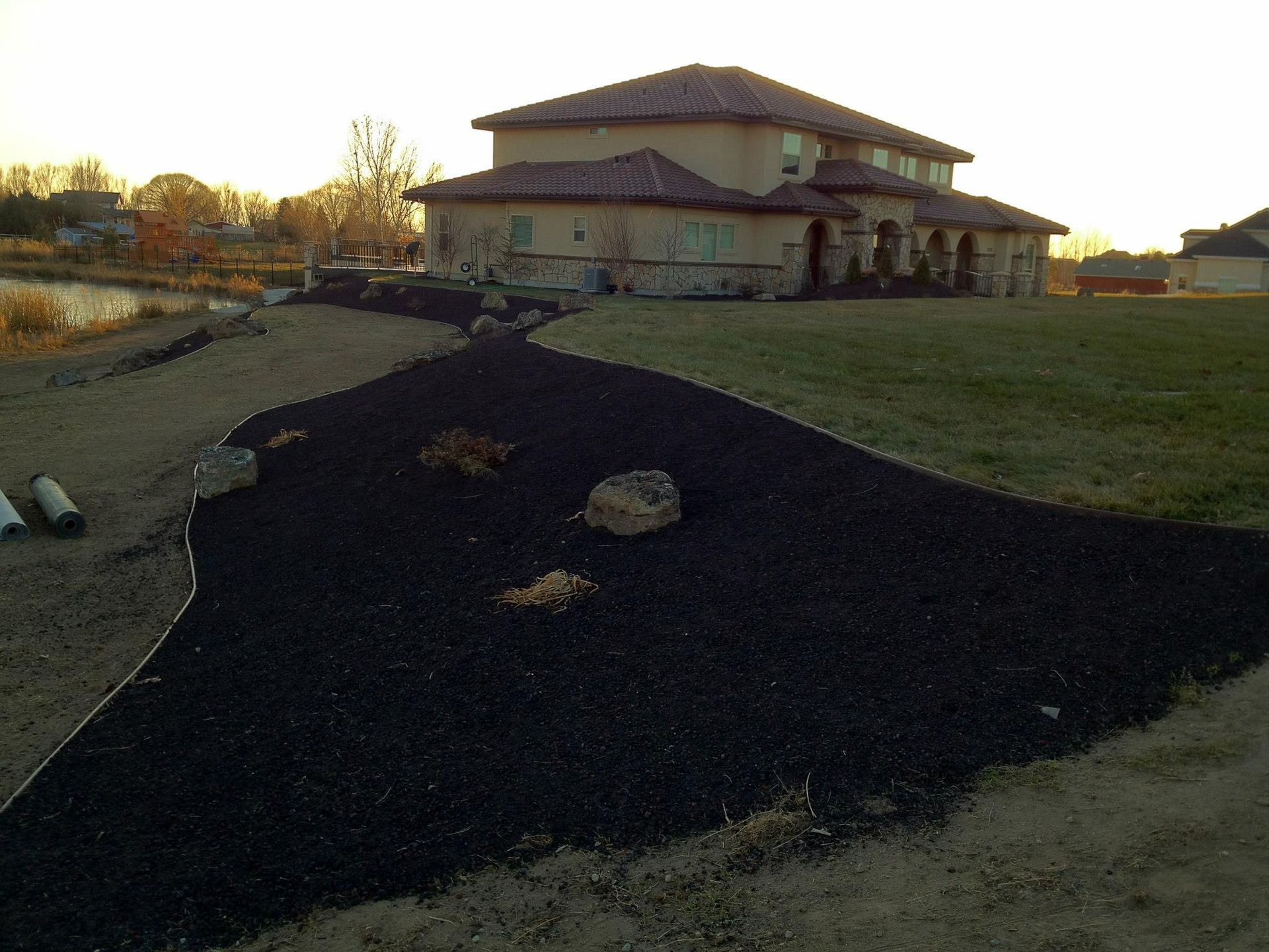 A landscaped yard featuring a curved patch of dark mulch bordered by rocks, with a large house in the background.