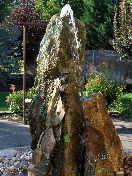 A tall, rugged, mossy rock fountain sprays a small jet of water from its peak into a bed of smooth river stones.