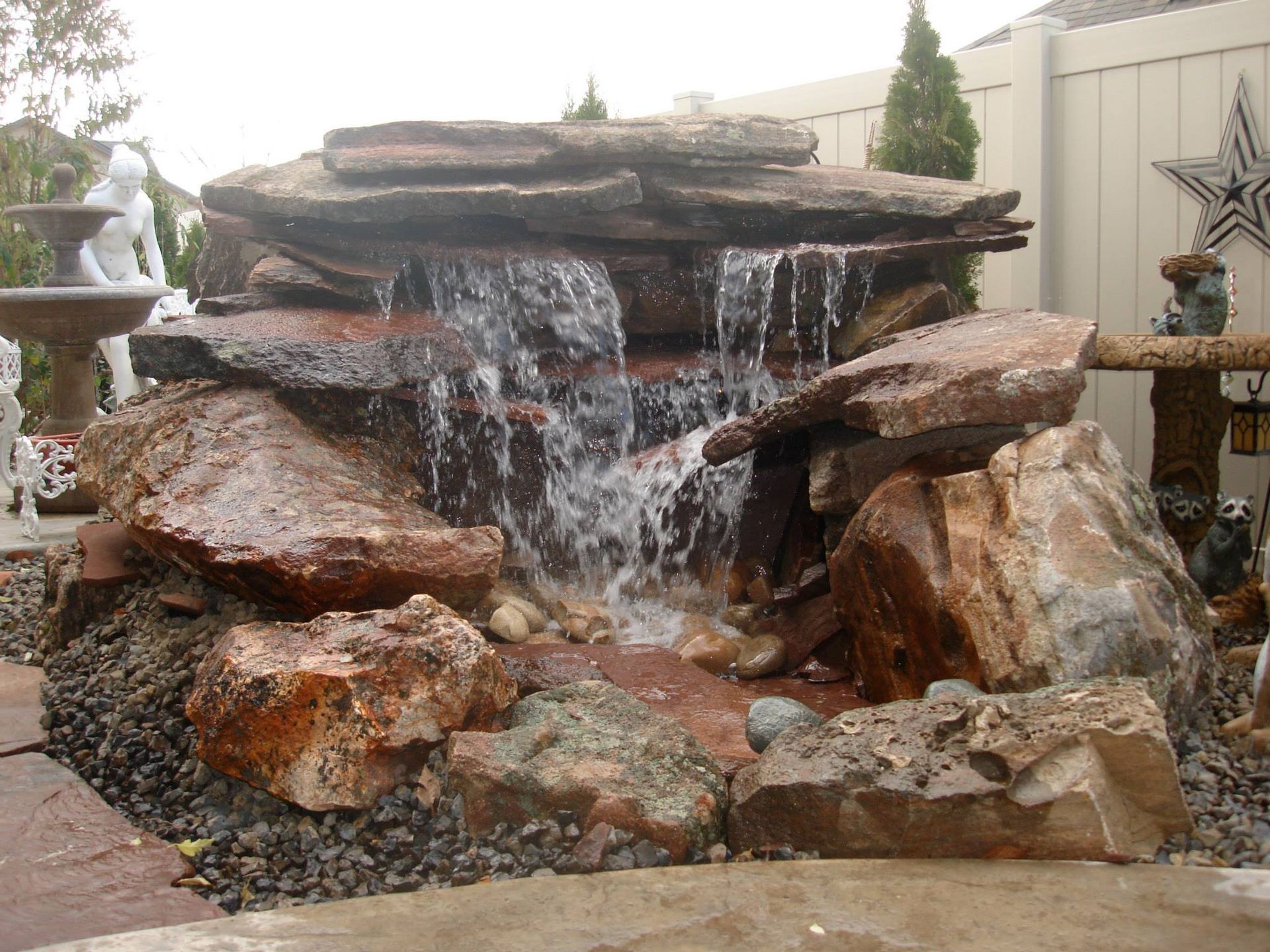 A backyard rock waterfall feature with water cascading over stacked natural stone slabs into a pebble-covered basin.