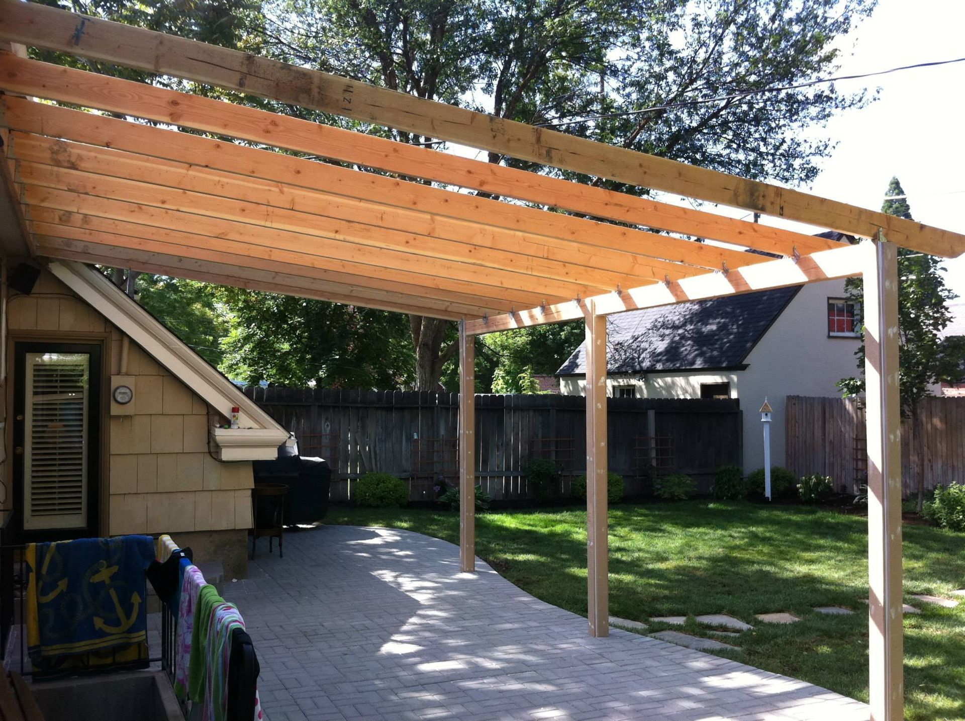 A wooden pergola frame under construction over a stone patio in a sunny backyard.