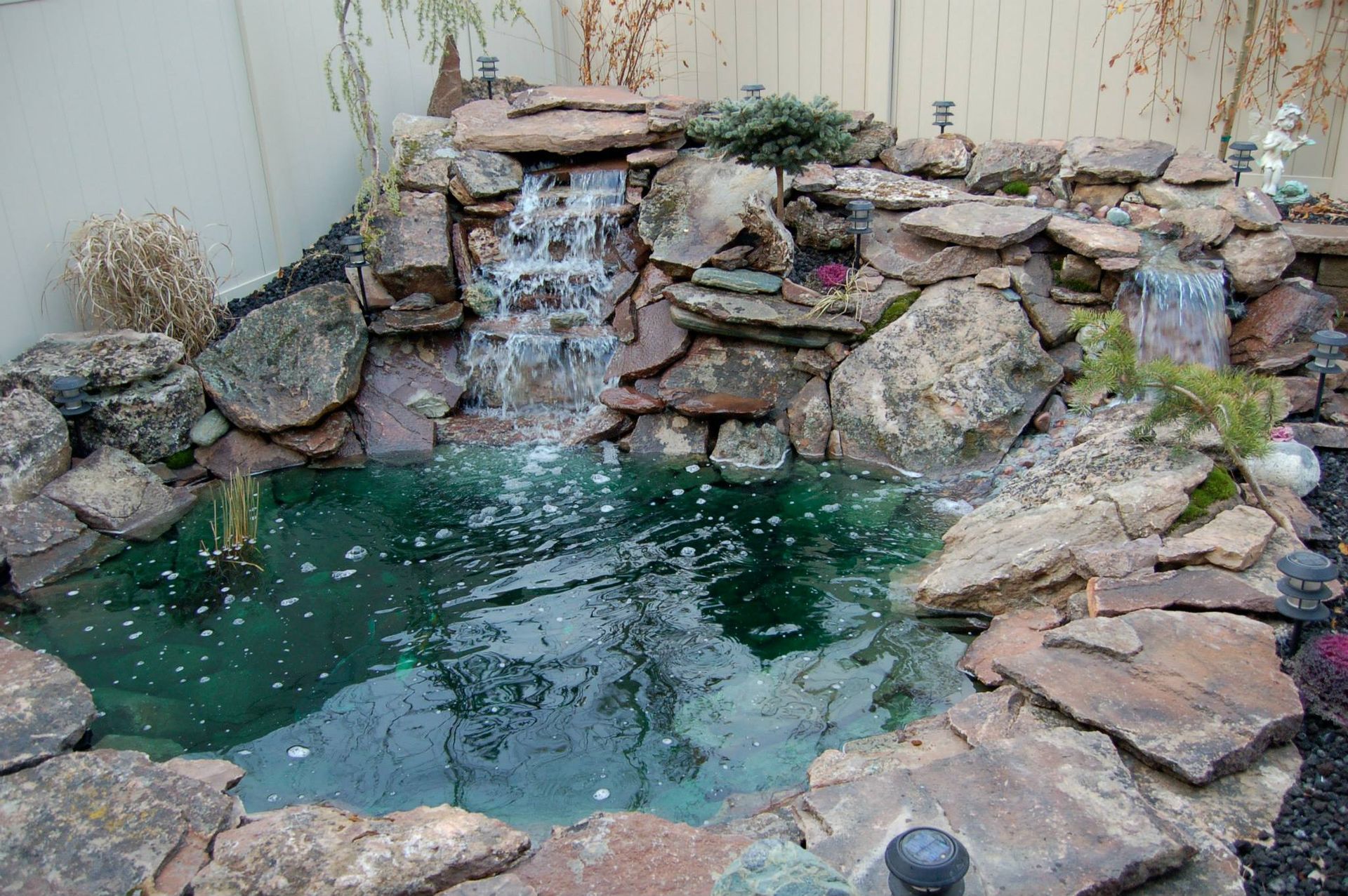 A backyard pond with rocky borders and two tiered waterfalls flowing into a clear, teal-colored pool.