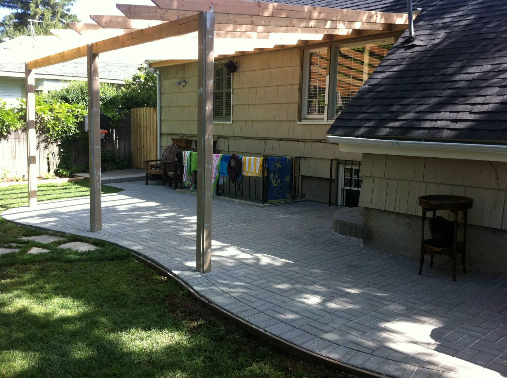 A wooden pergola stands over a paved stone patio next to a house with beige siding and a dark shingled roof.