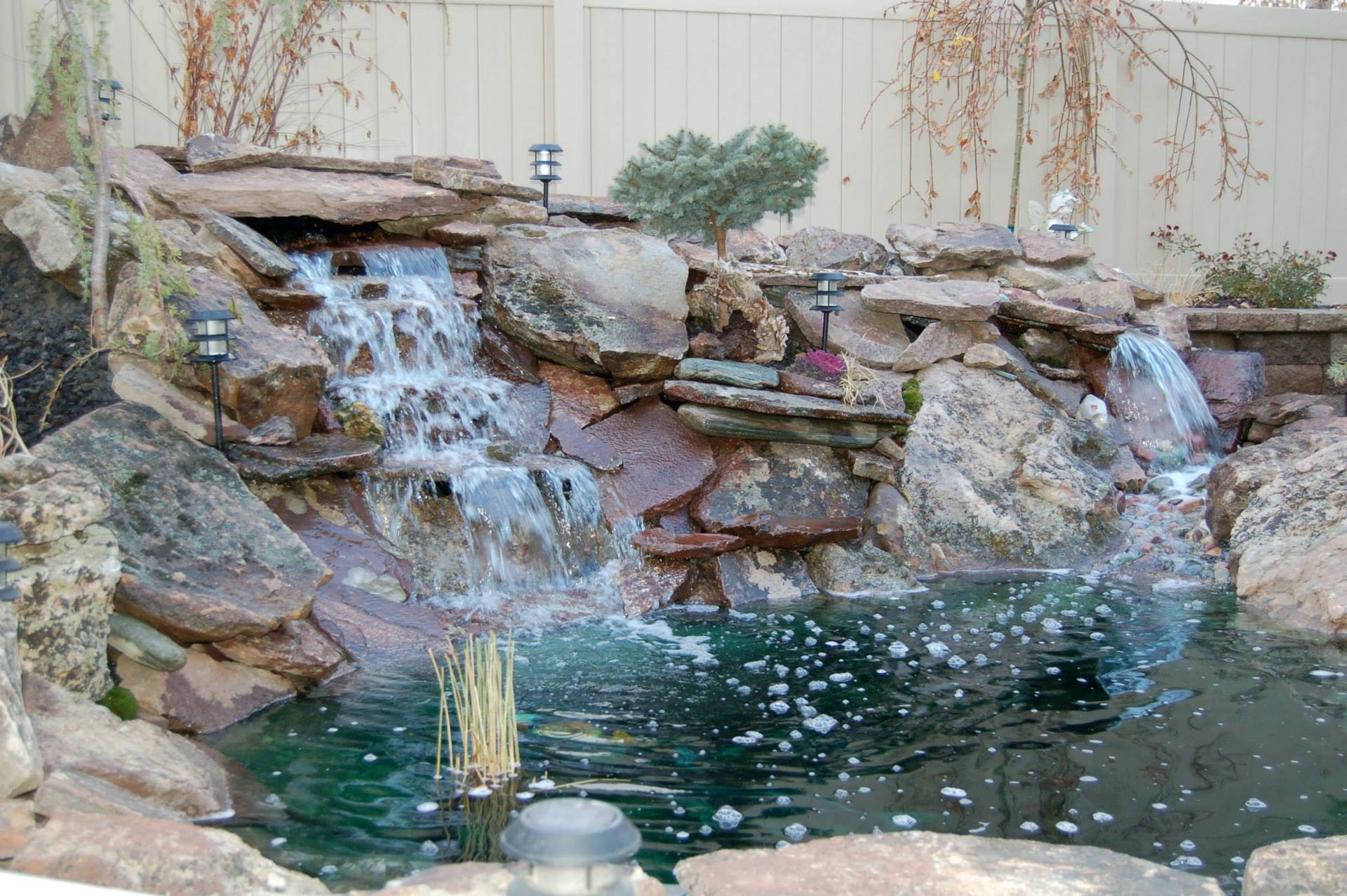 A two-tier backyard waterfall flows over natural tan and grey rocks into a dark, small koi pond.
