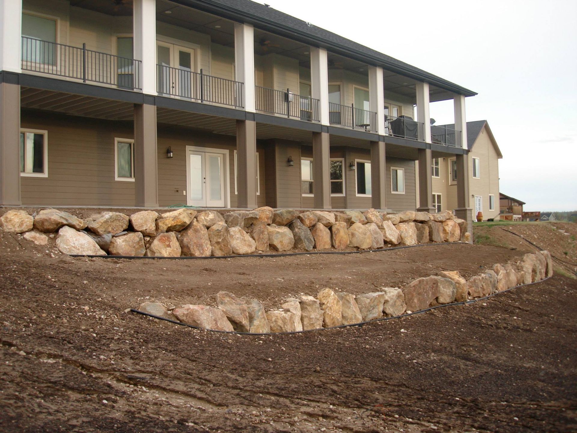 A tiered stone retaining wall landscape leading to the back porch of a beige multi-story house.