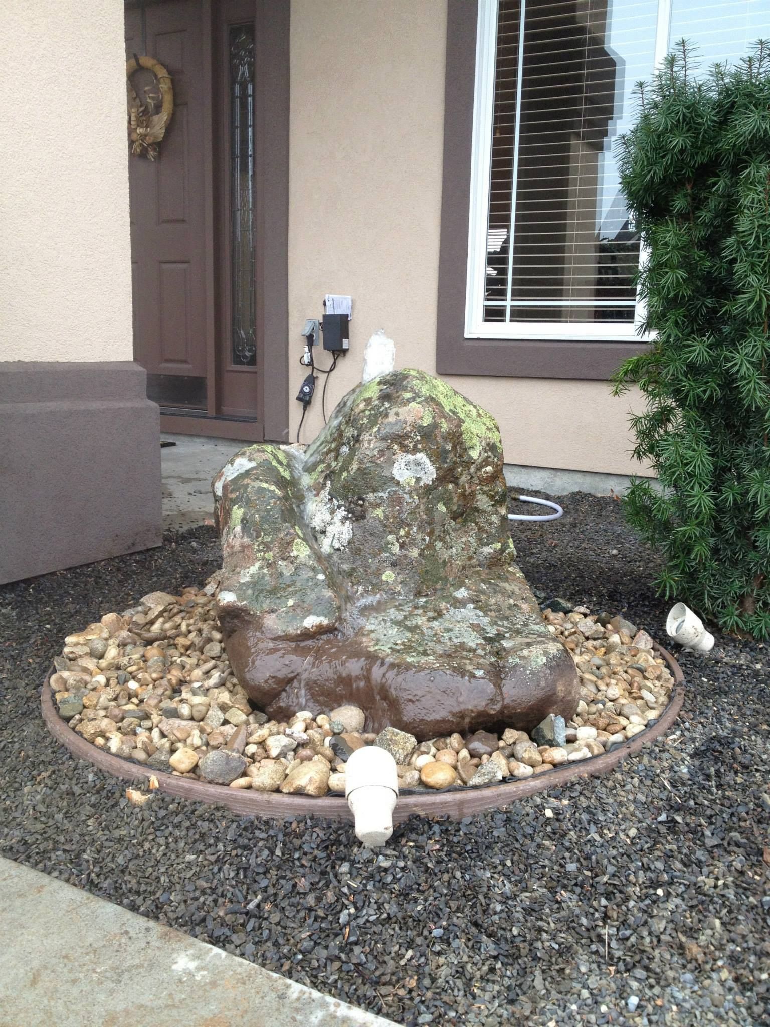 A large, wet, moss-covered decorative rock fountain sits in a gravel bed in front of a house entrance.