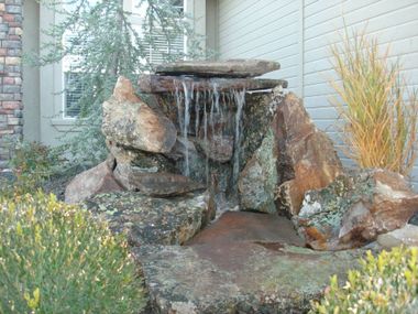 A stone garden waterfall feature with water trickling down natural rocks, surrounded by greenery in front of a house.