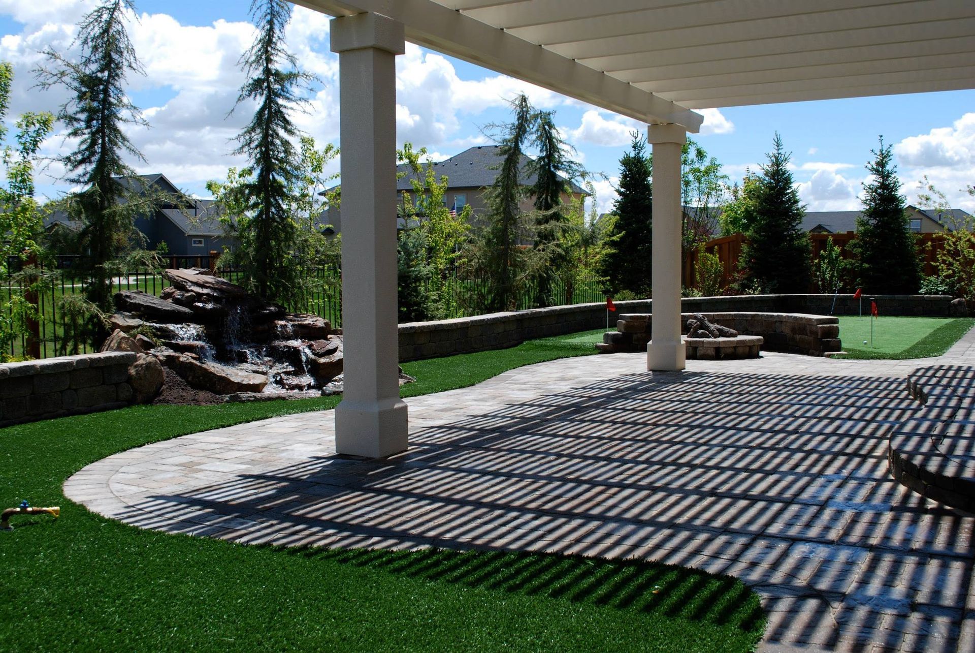 A paved patio with a pergola overhead, featuring shadow patterns on the ground, a stone water feature, and a grassy yard.