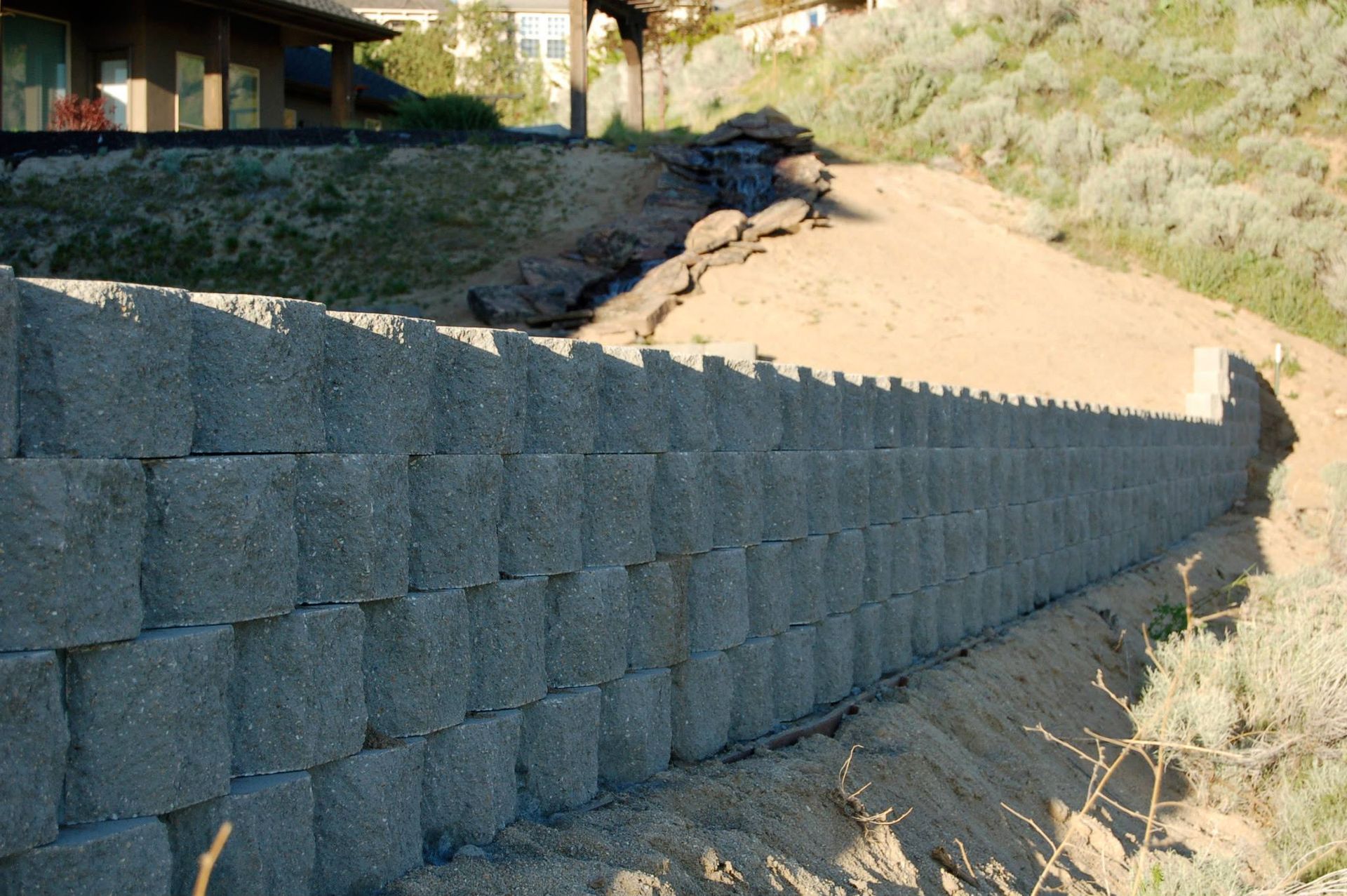 A long, newly constructed gray concrete block retaining wall sits in front of a sandy, sloped hillside.