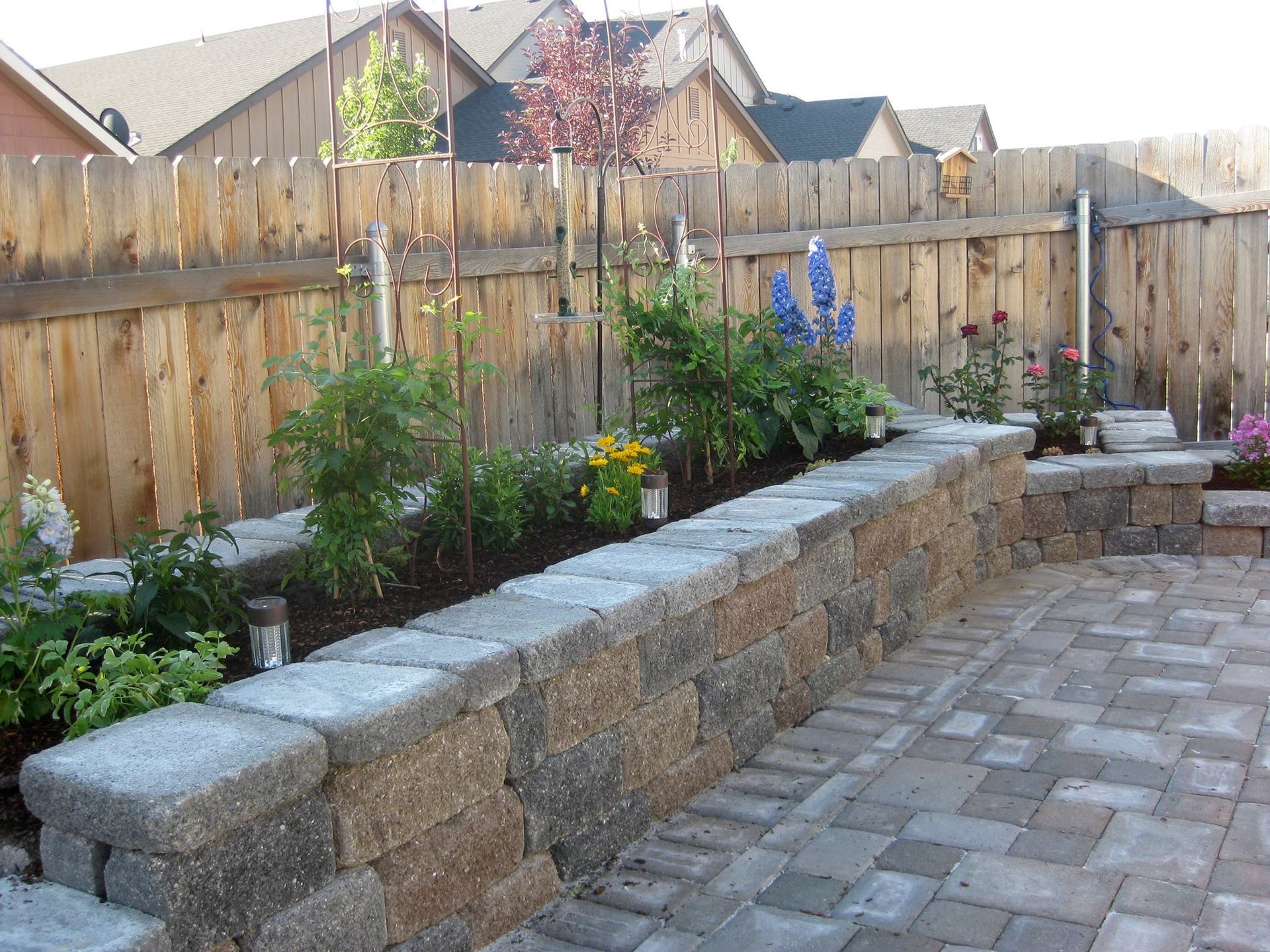 A raised stone garden bed filled with green plants and blue flowers sits on a patio in front of a wooden fence.