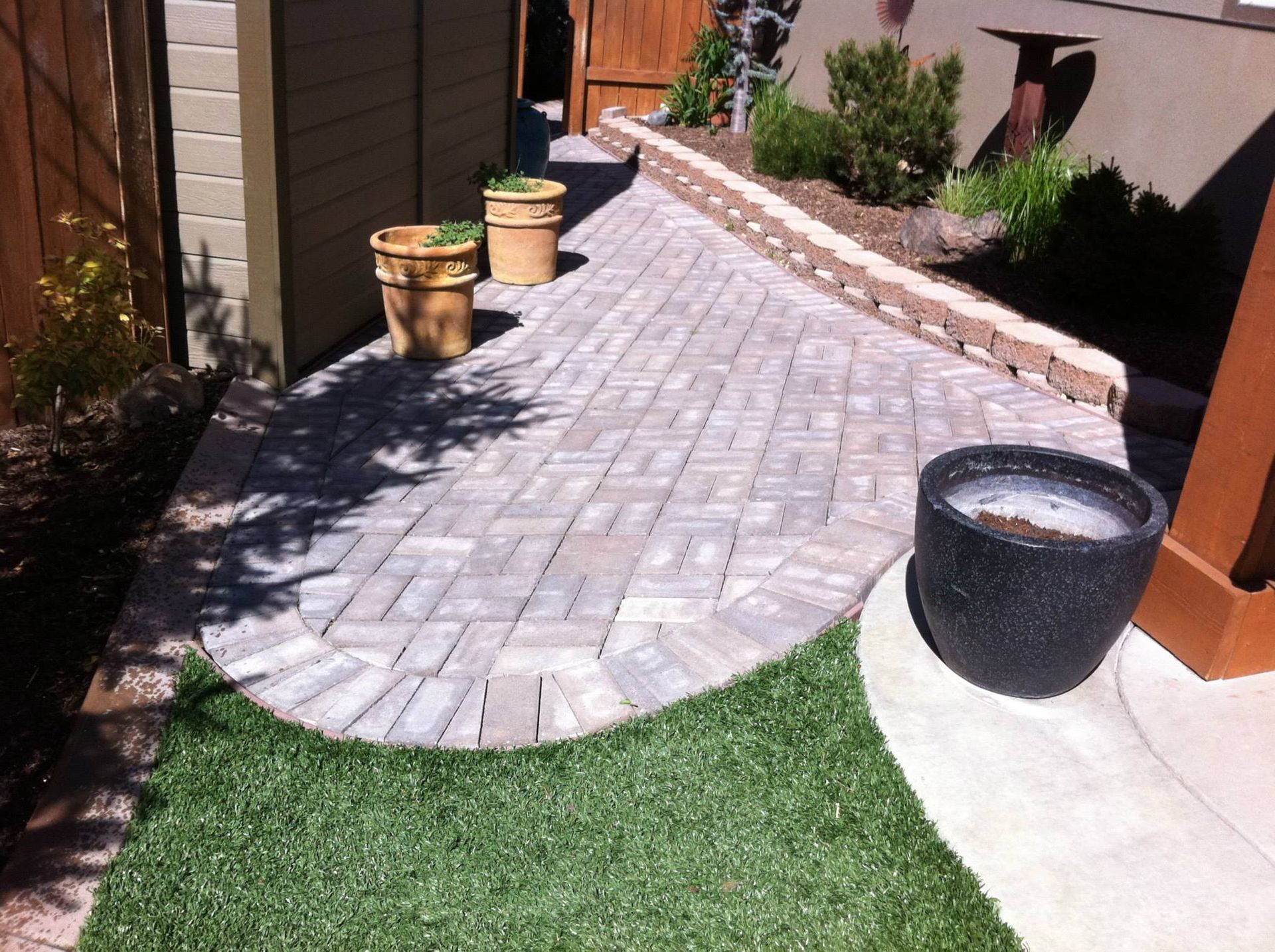 A paved brick walkway curves through a backyard, bordered by artificial grass, potted plants, and a small stone retaining wall.