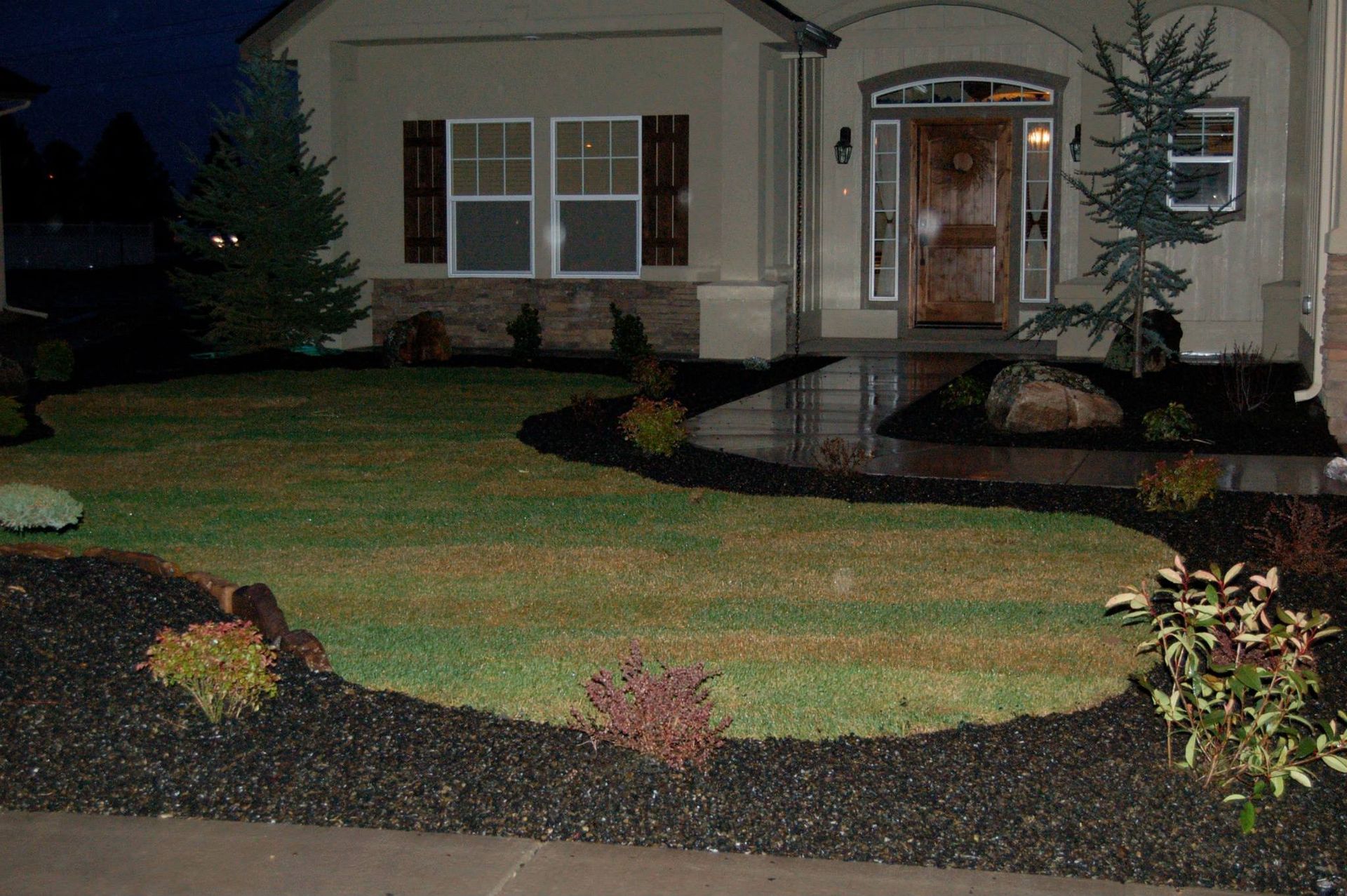 A front yard at night with a manicured lawn, dark mulch flower beds, small shrubs, and two evergreen trees near a house.