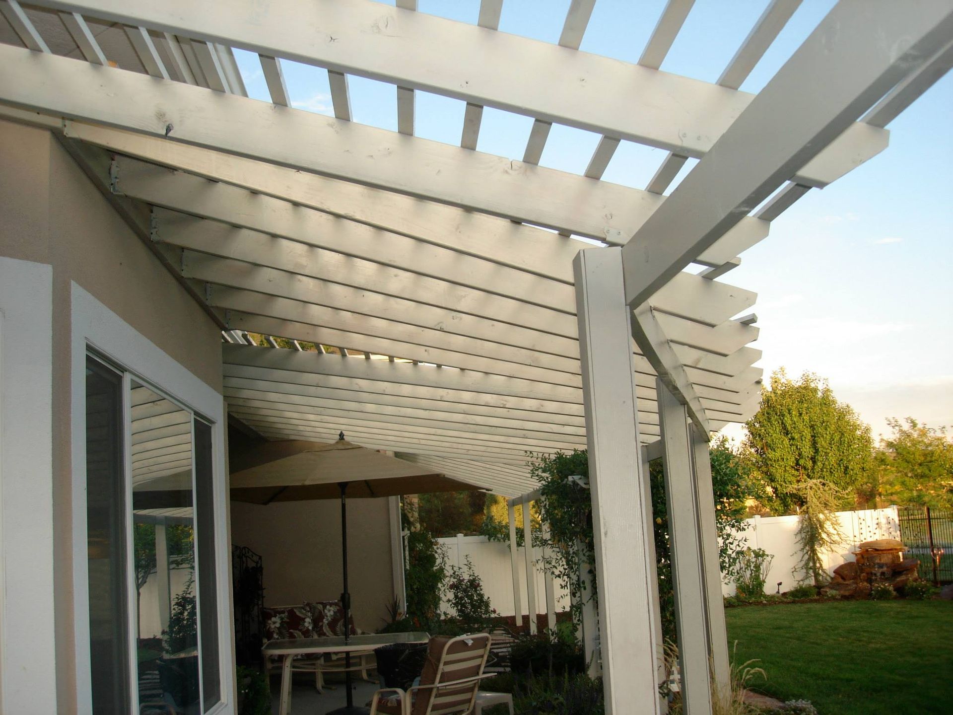 A white wooden pergola extends from a house, shading a patio with chairs and an umbrella on a sunny day.