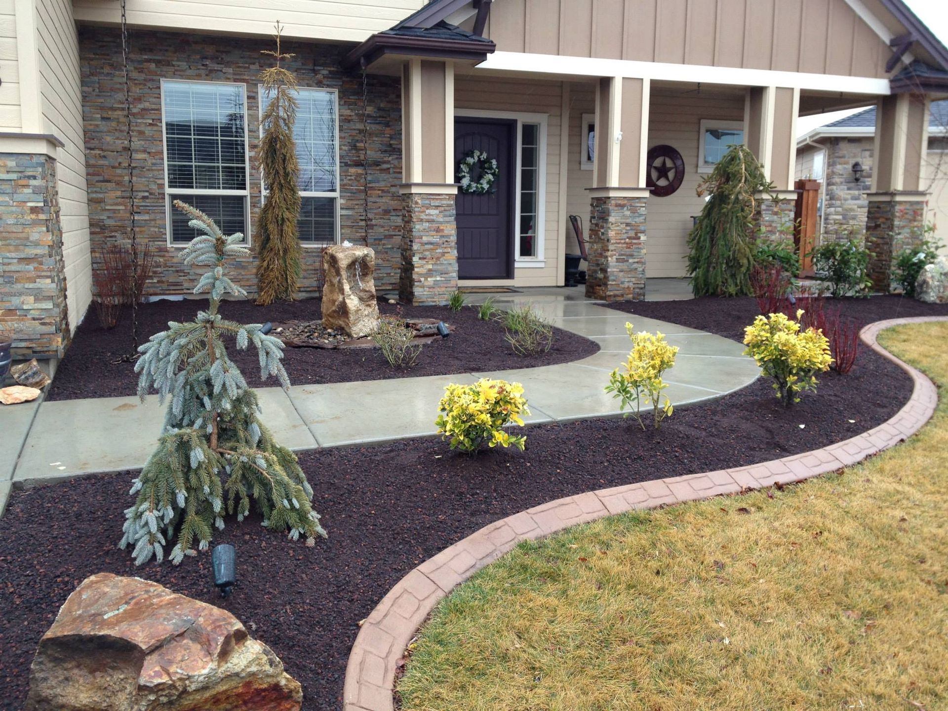 A suburban house front yard with a curved stone walkway, dark mulch, decorative boulders, and small shrubs.