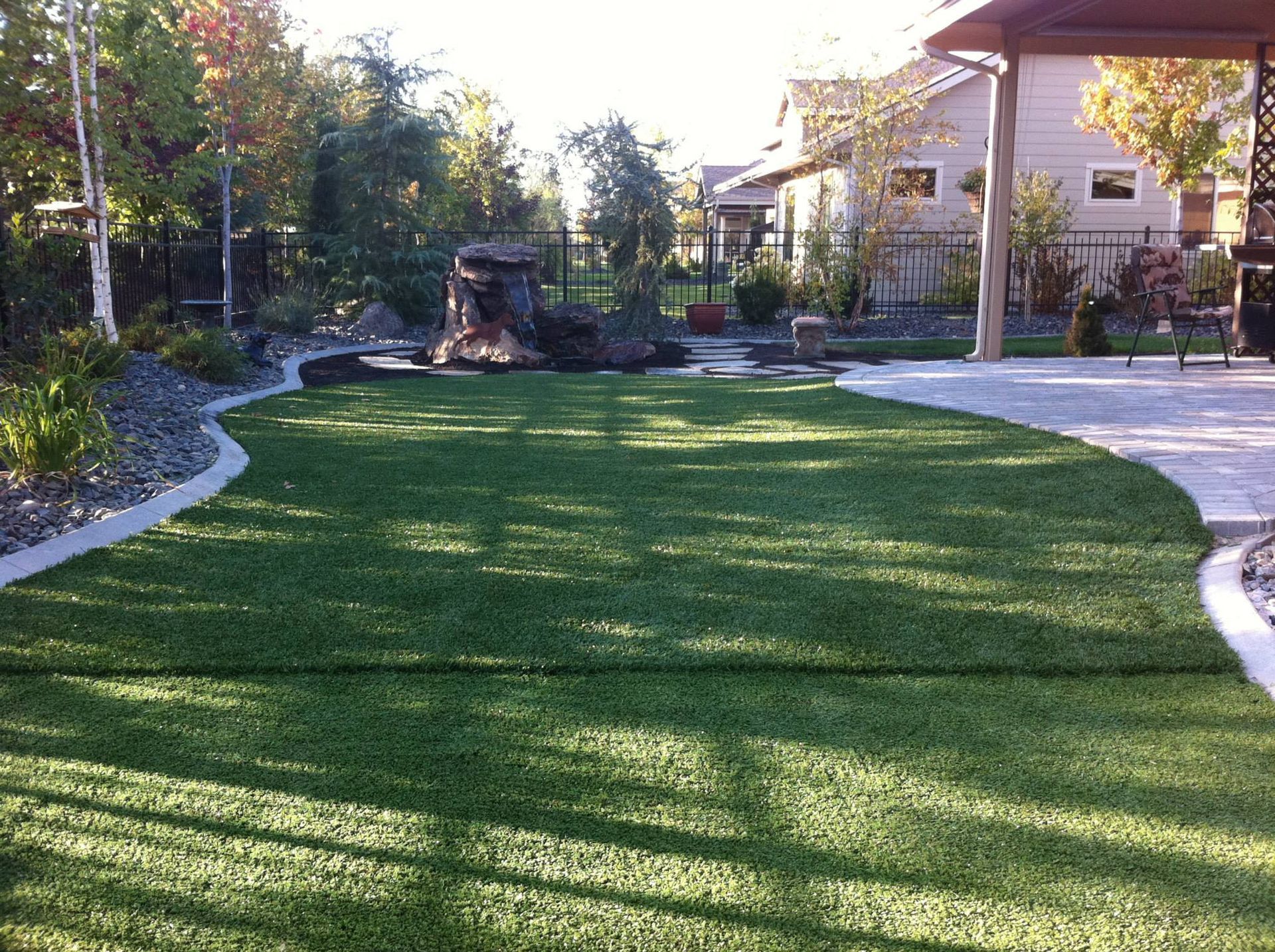 A landscaped backyard with a green lawn bordered by a stone edge, a central rock feature, and a patio under a canopy.