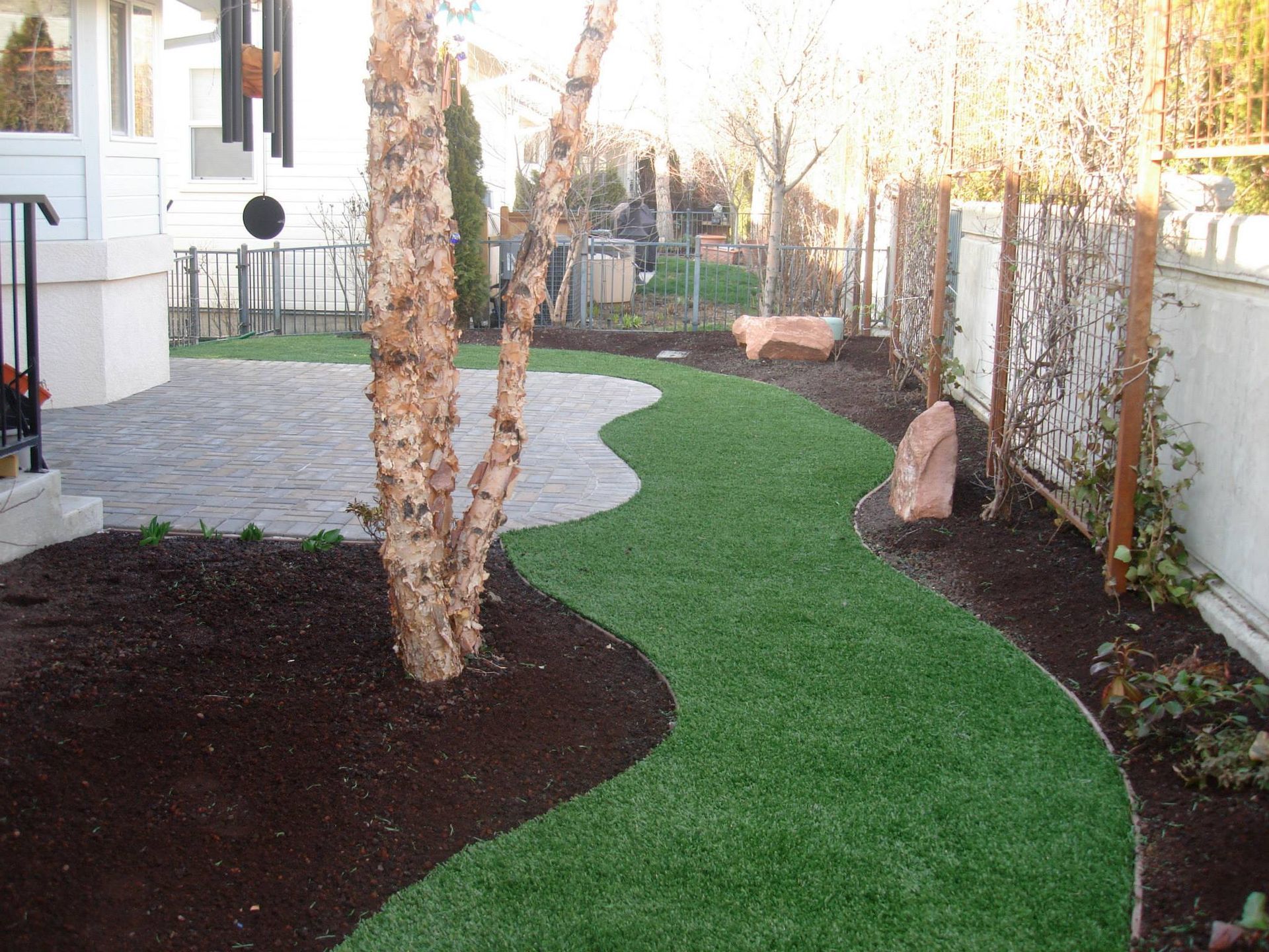 A winding path of green turf runs between a stone patio and a mulched garden bed with trees and a white fence.