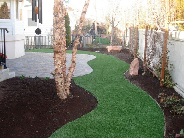 A winding path of green turf runs between a stone patio and a mulched garden bed with trees and a white fence.