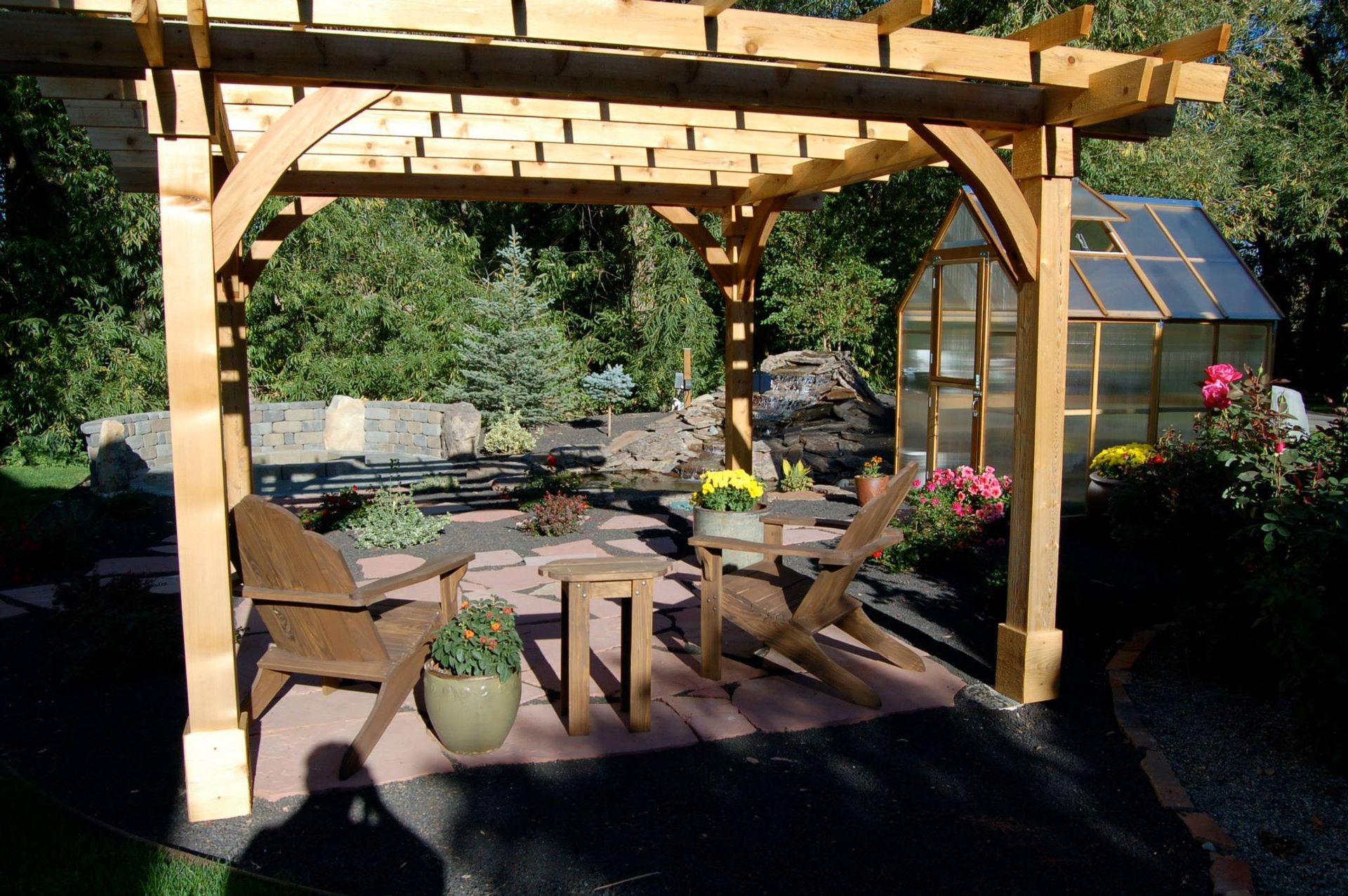 Two wooden Adirondack chairs and a small table sit under a pergola on a stone patio, with a greenhouse in the background.