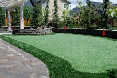 A backyard putting green with two red flags, set against a stone retaining wall, patio, and landscaping.