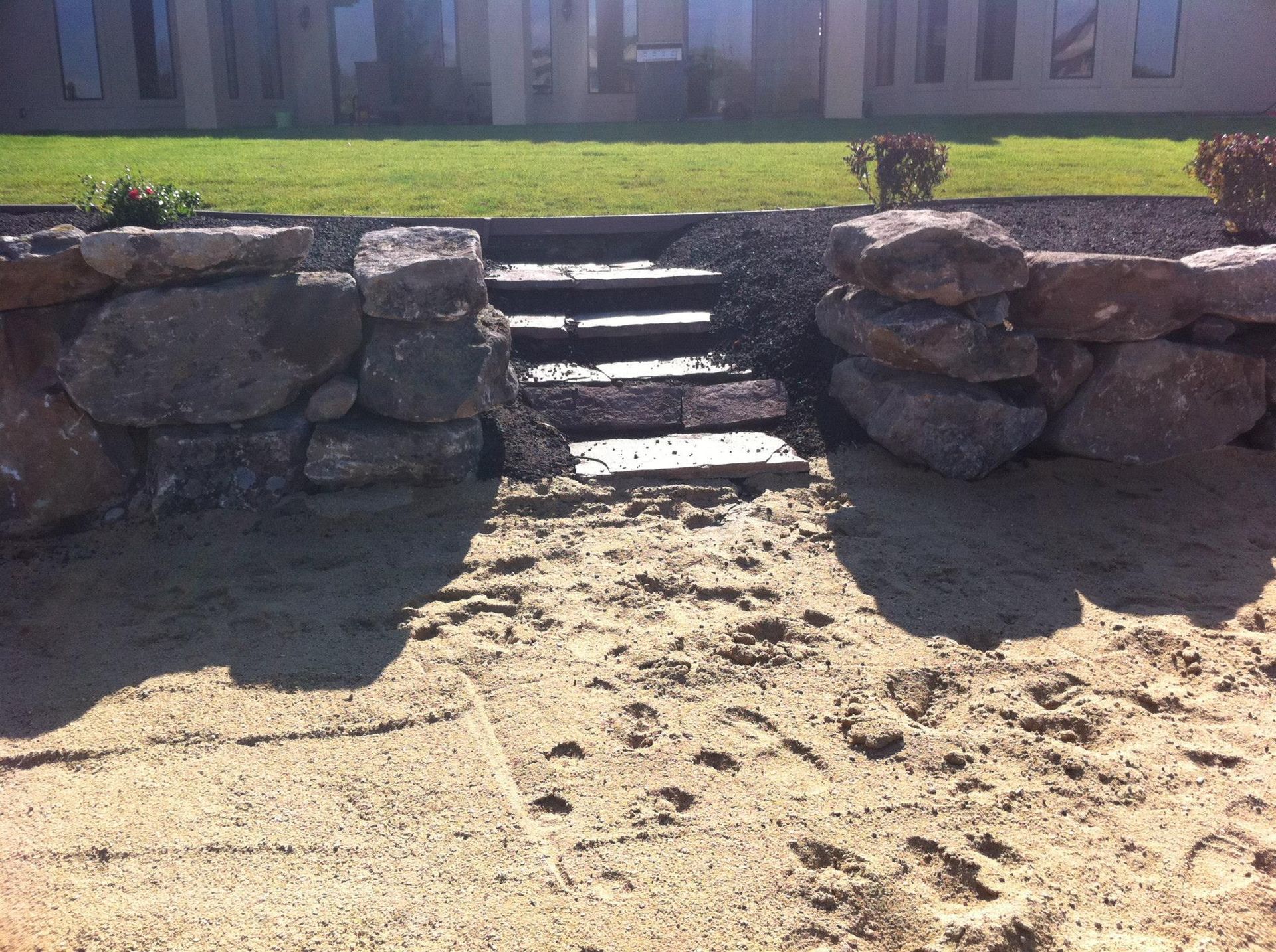 Stone steps set into a low boulder retaining wall, leading from a sandy area up to a lawn in front of a house.