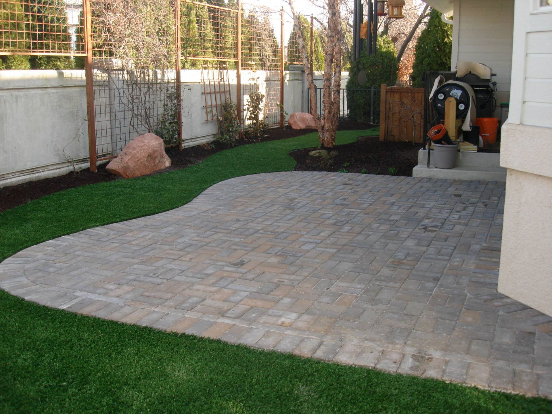 A stone patio with a curved edge transitions into a lush green lawn, next to a house wall and a backyard fence.