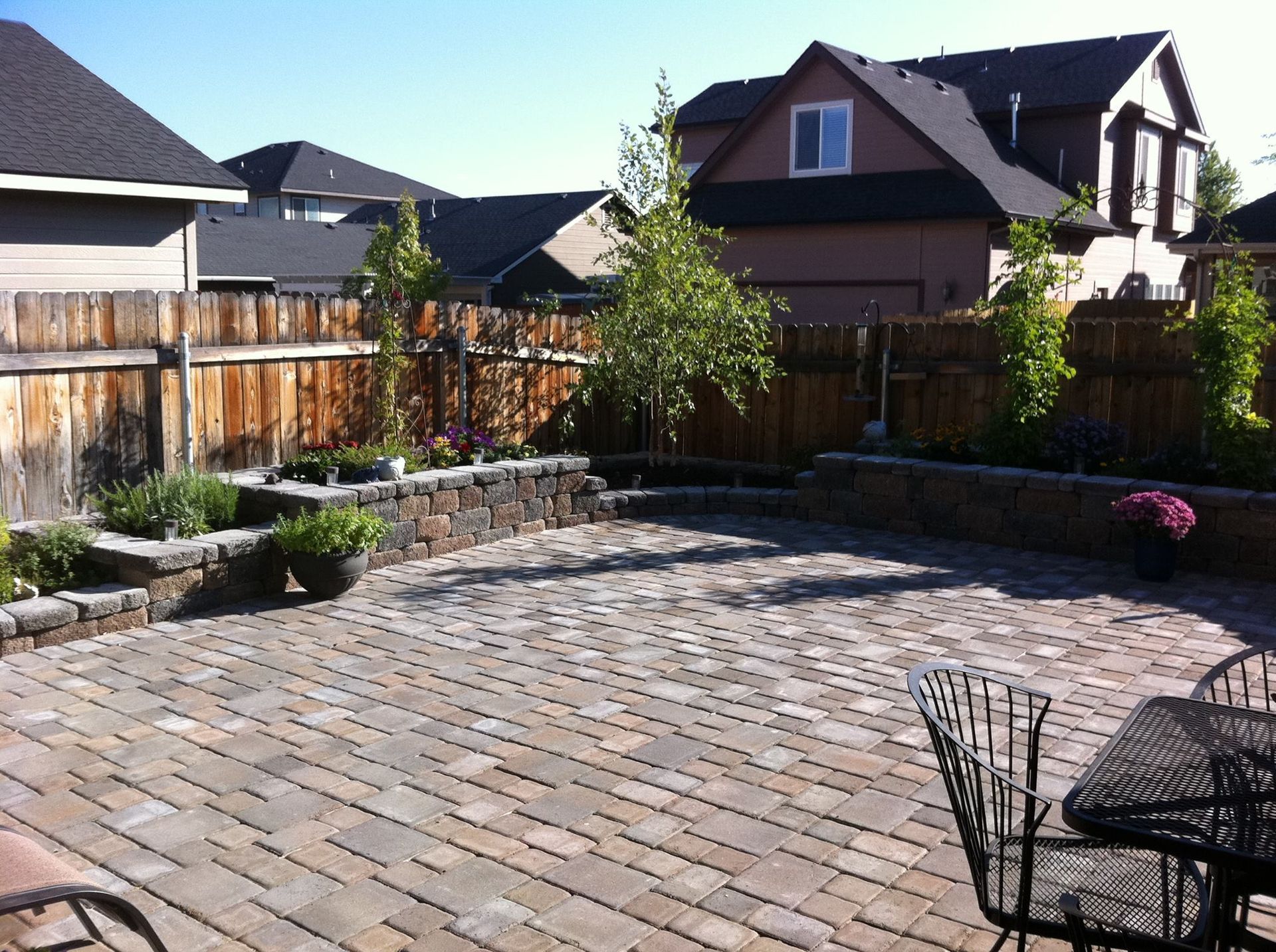 A stone-paved backyard patio featuring tiered garden beds, wooden fencing, and outdoor furniture under a sunny sky.
