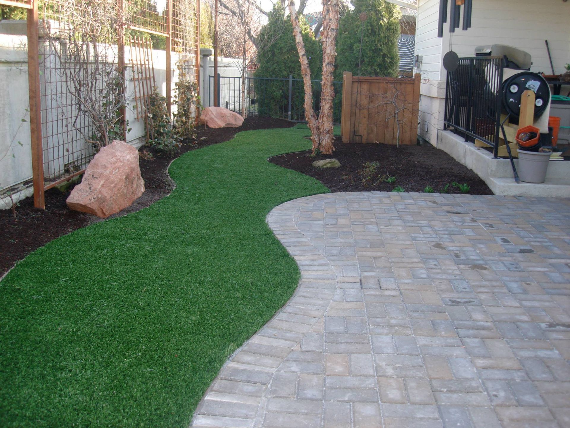 A curved path of artificial green grass alongside a stone paver patio in a residential backyard with garden beds.