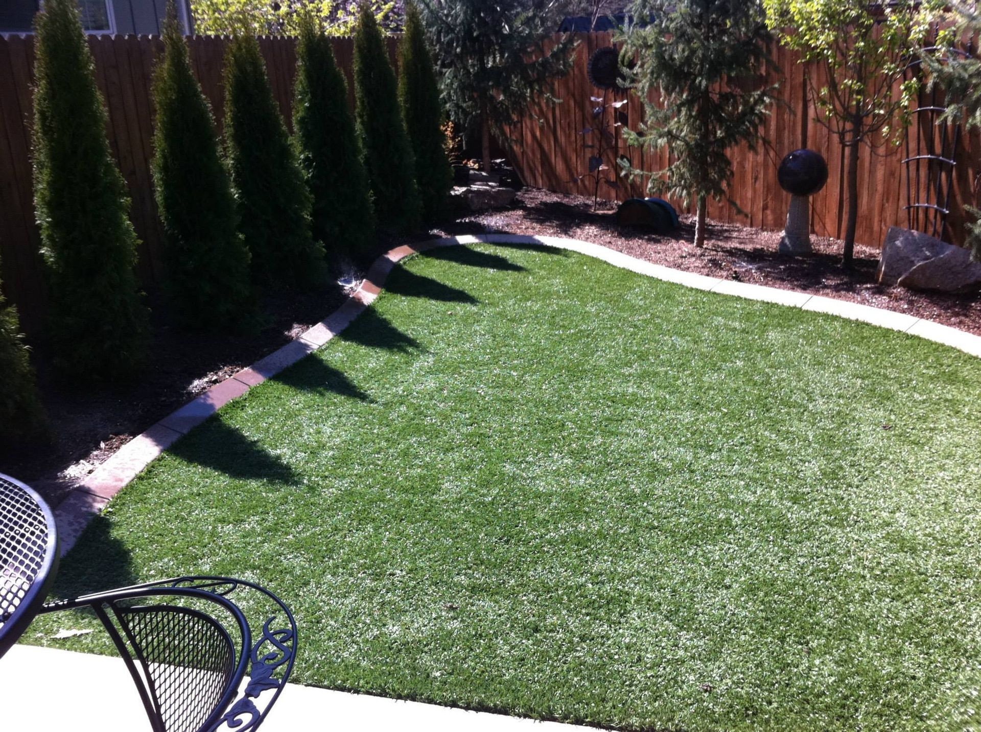 A small, lush green synthetic lawn bordered by pavers, with a row of evergreen trees and a wood fence in the background.